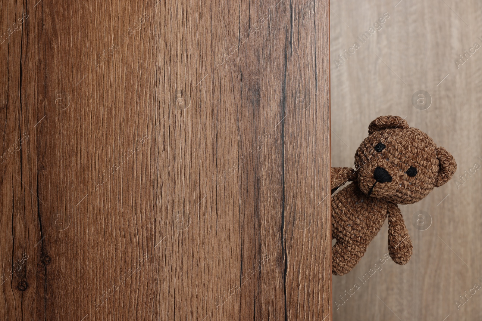 Photo of Cute knitted bear peeking out of wooden door, space for text