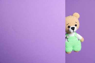 Photo of Cute knitted bear peeking out from color card on violet background, space for text