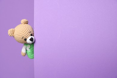 Photo of Cute knitted bear peeking out from color card on violet background, space for text