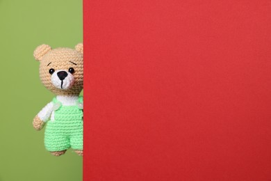 Photo of Cute knitted bear peeking out from red card on green background, space for text