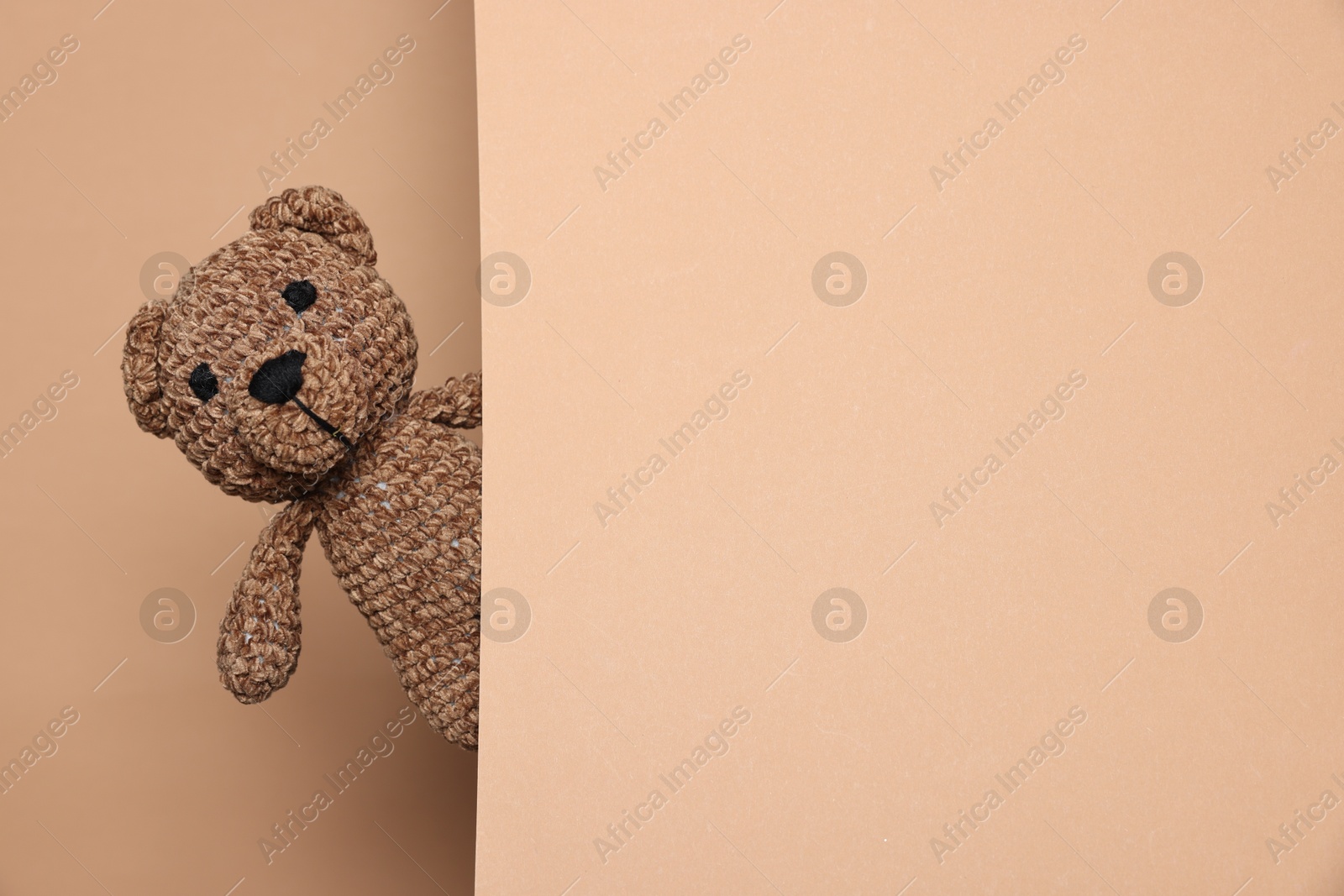 Photo of Cute knitted bear peeking out from beige card on brown background, space for text