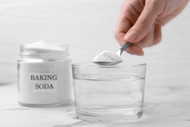 Photo of Woman adding baking soda to glass of water at white marble table, closeup