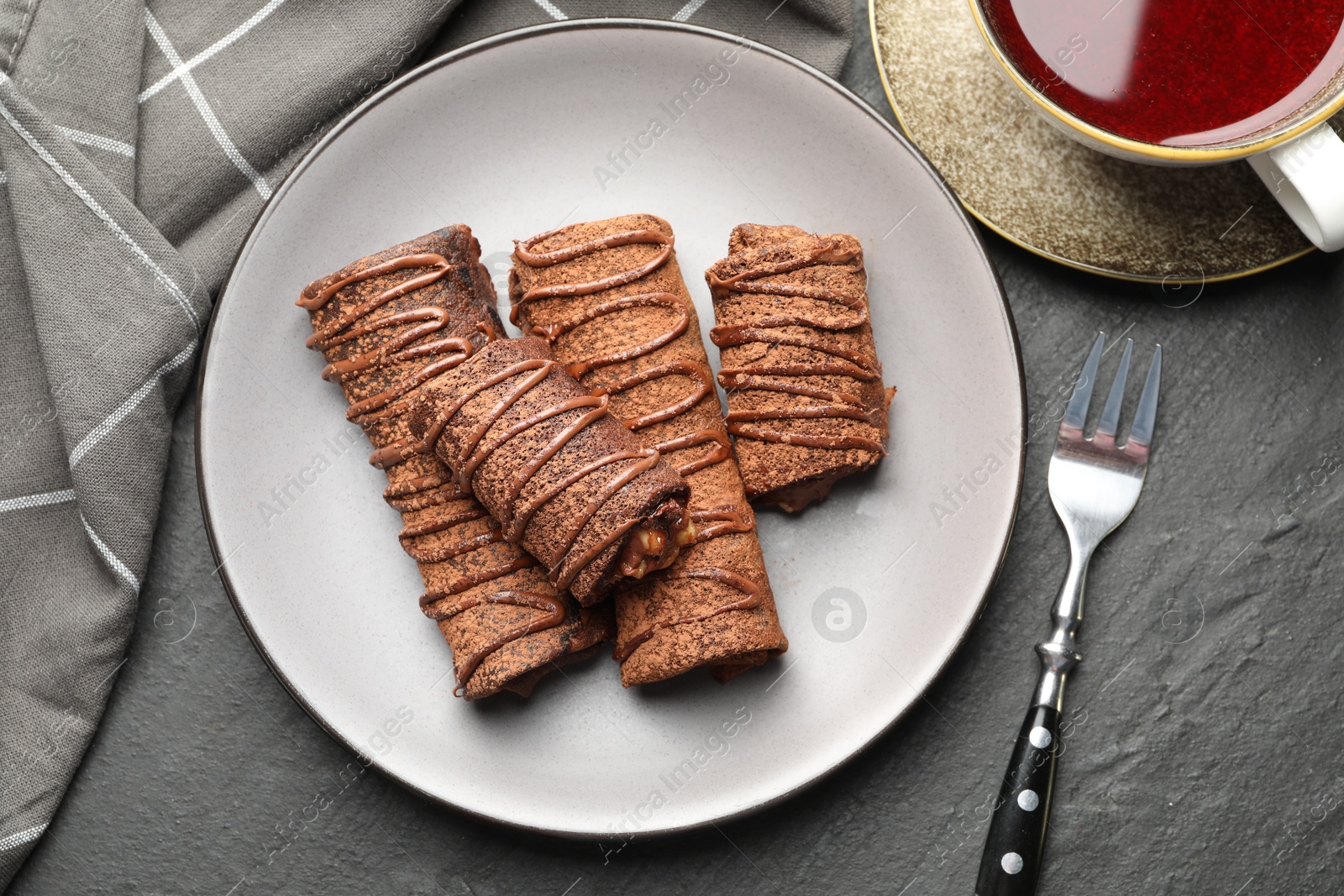 Photo of Delicious chocolate crepes with sweet paste, nuts and tea on black table, flat lay