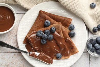 Photo of Delicious chocolate crepes with sweet paste and blueberries on light tiled table, flat lay