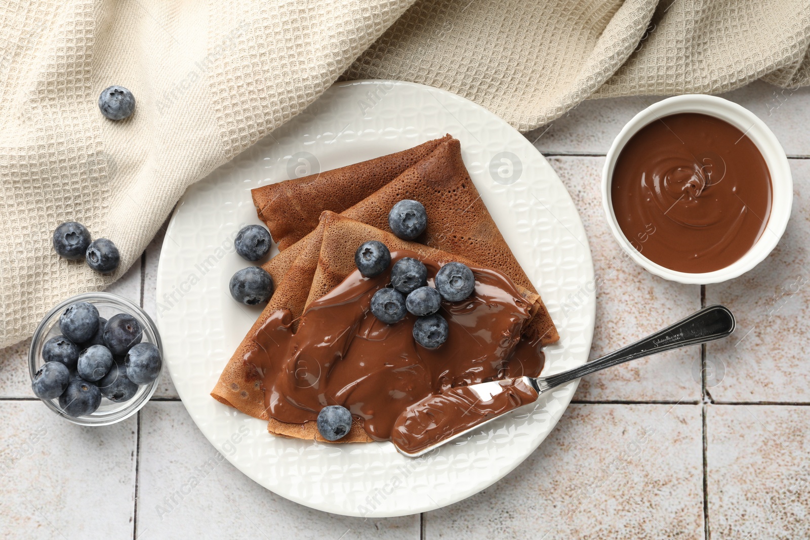 Photo of Delicious chocolate crepes with sweet paste and blueberries on light tiled table, flat lay