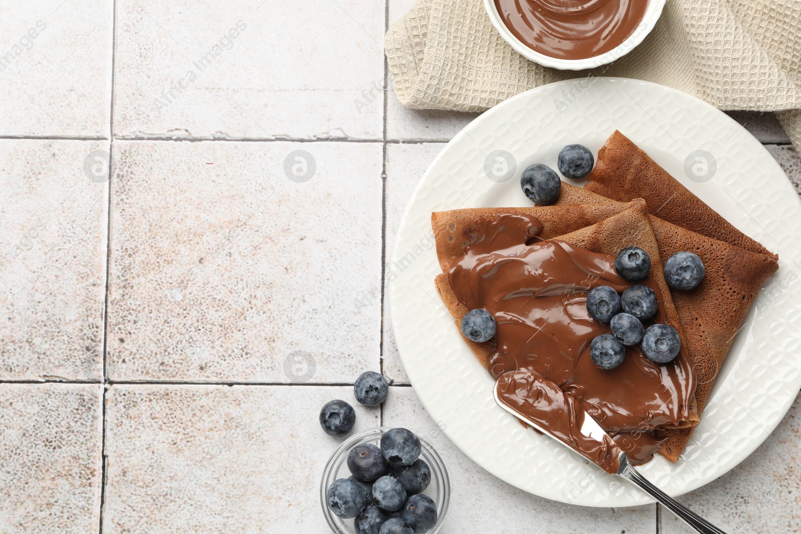 Photo of Delicious chocolate crepes with sweet paste and blueberries on light tiled table, flat lay. Space for text