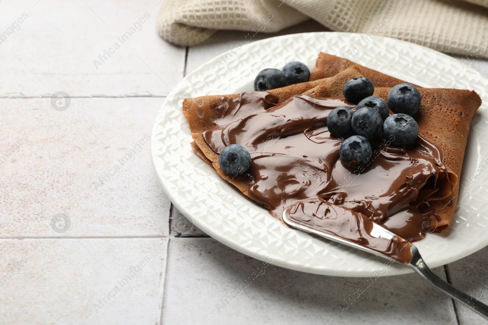 Photo of Delicious chocolate crepes with sweet paste and blueberries on light tiled table, closeup. Space for text