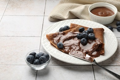 Photo of Delicious chocolate crepes with sweet paste and blueberries on light tiled table, closeup. Space for text