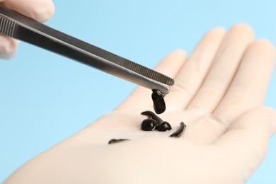 Photo of Woman holding medicinal leech with tweezers on light blue background, closeup