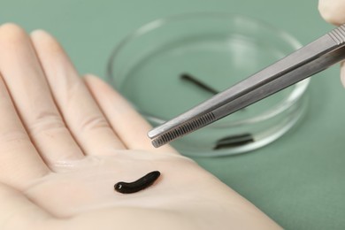 Photo of Woman holding medicinal leech and tweezers on green background, closeup