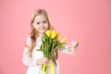 Photo of Cute little girl with yellow tulips and decorative bunny on pink background. Easter celebration