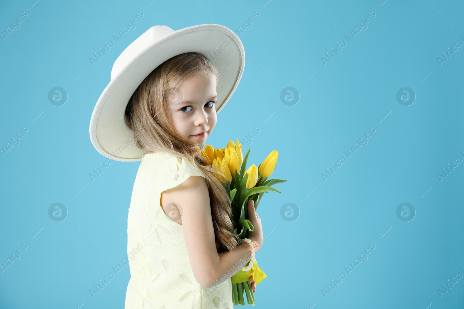 Photo of Cute little girl with beautiful yellow tulips on light blue background, space for text. Spring season