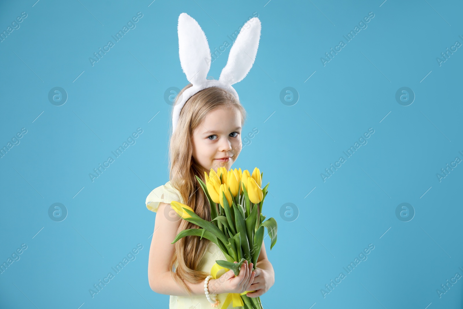 Photo of Cute little girl with bunny ears and tulips on light blue background. Easter celebration