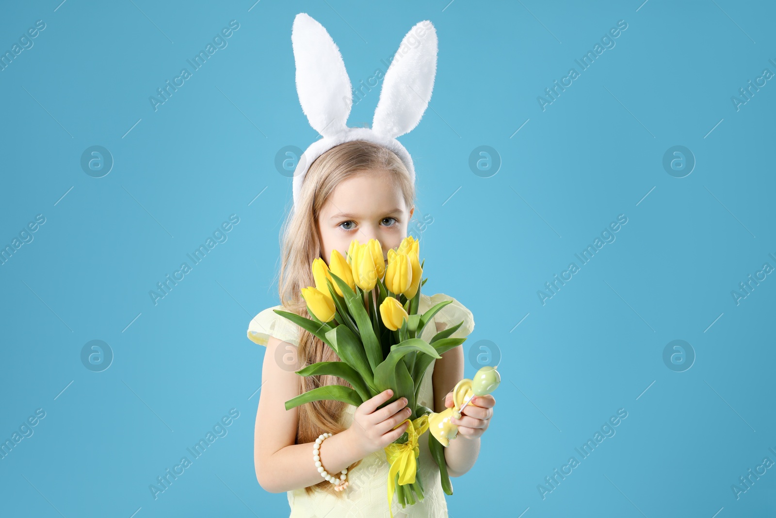 Photo of Cute little girl with bunny ears and tulips on light blue background. Easter celebration