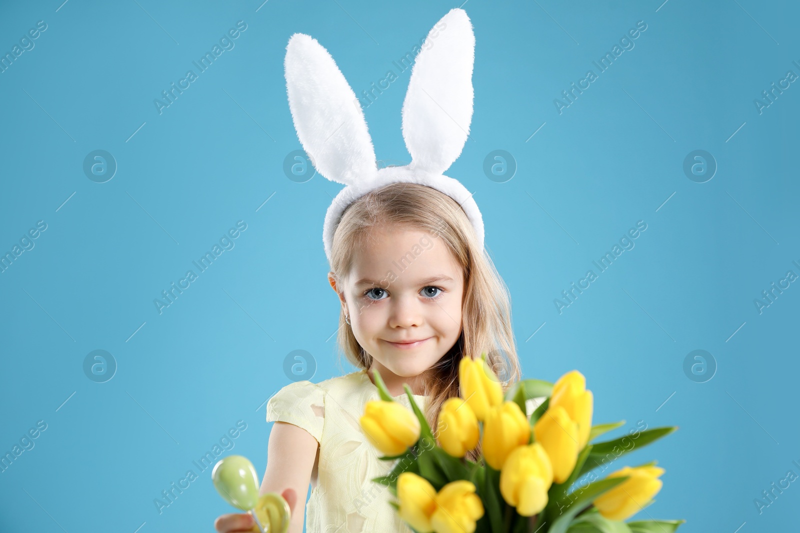 Photo of Cute little girl with bunny ears and tulips on light blue background. Easter celebration