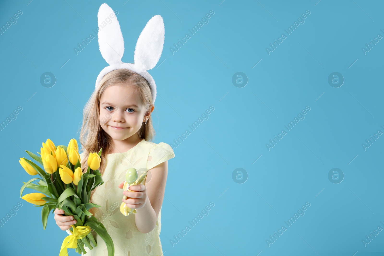 Photo of Cute little girl with bunny ears and tulips on light blue background, space for text. Easter celebration