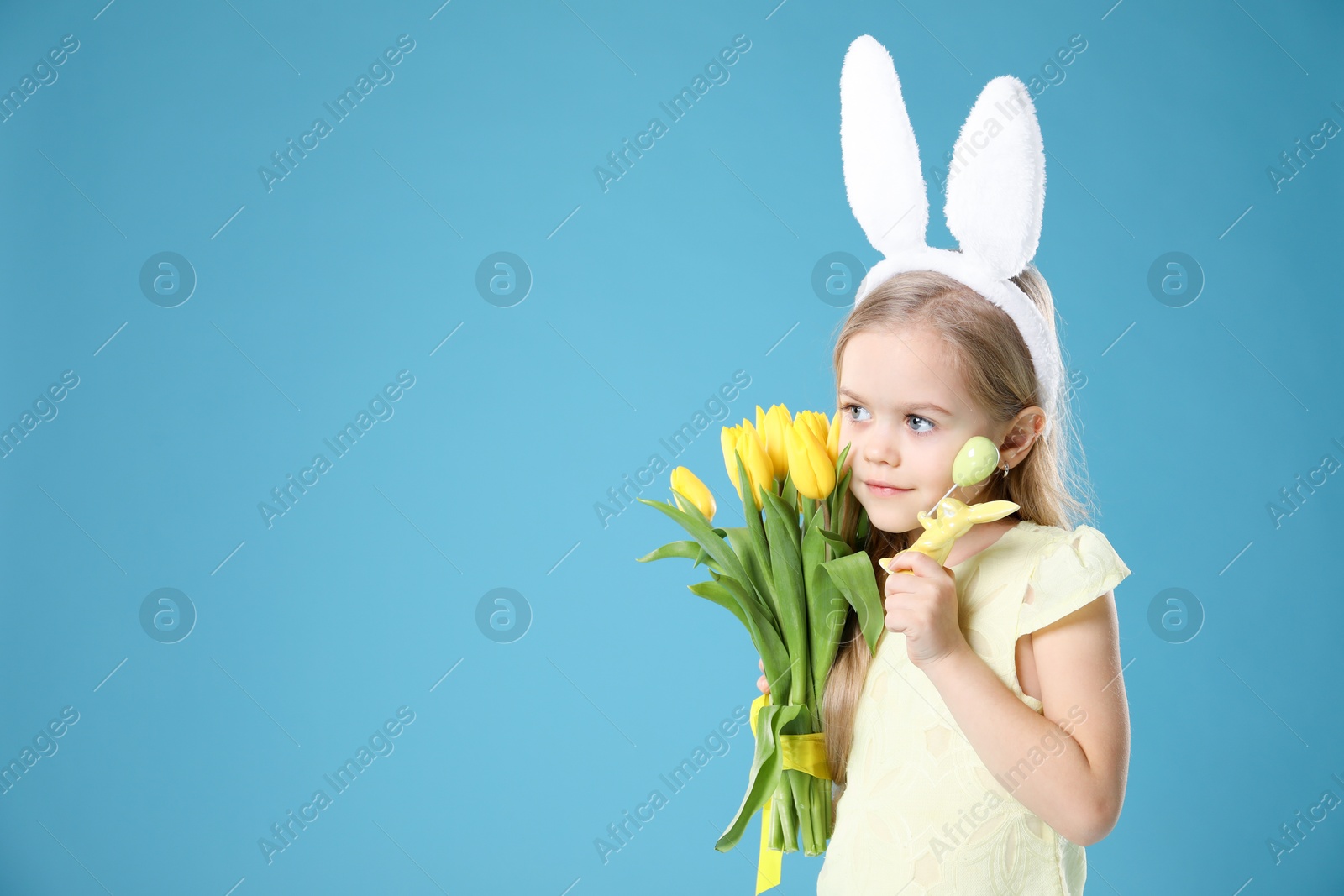 Photo of Cute little girl with bunny ears and tulips on light blue background, space for text. Easter celebration