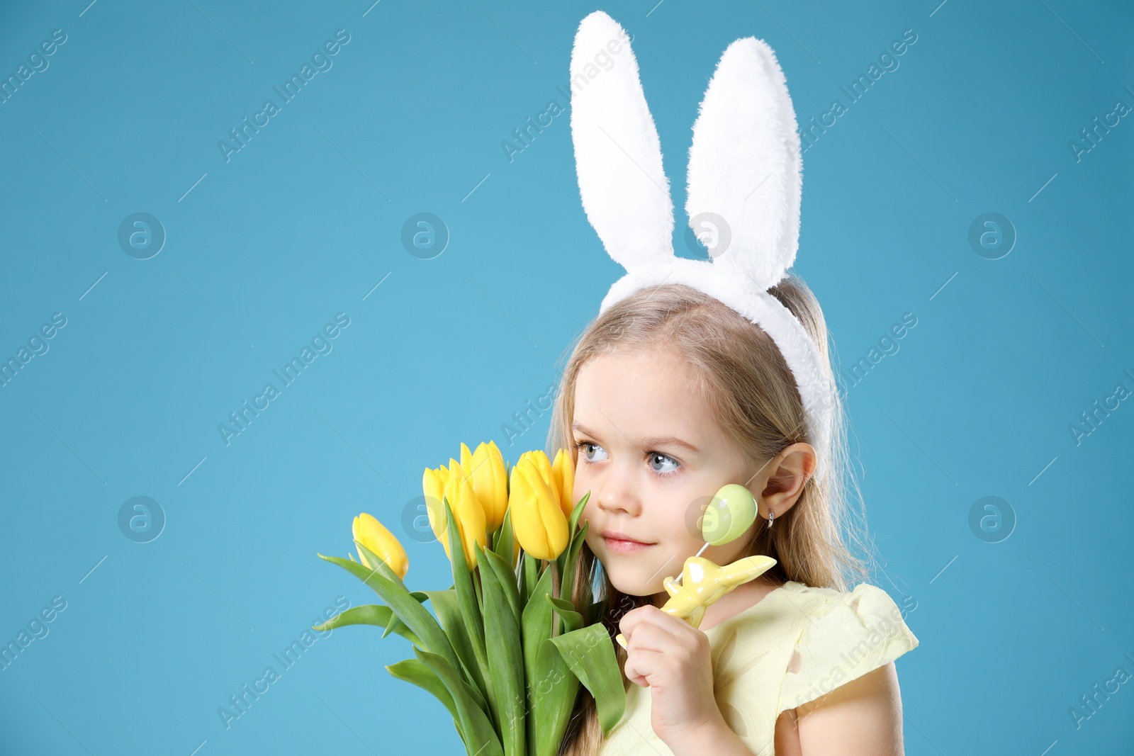 Photo of Cute little girl with bunny ears and tulips on light blue background. Easter celebration