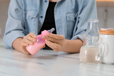 Photo of Mother making baby formula in feeding bottle at table indoors, closeup