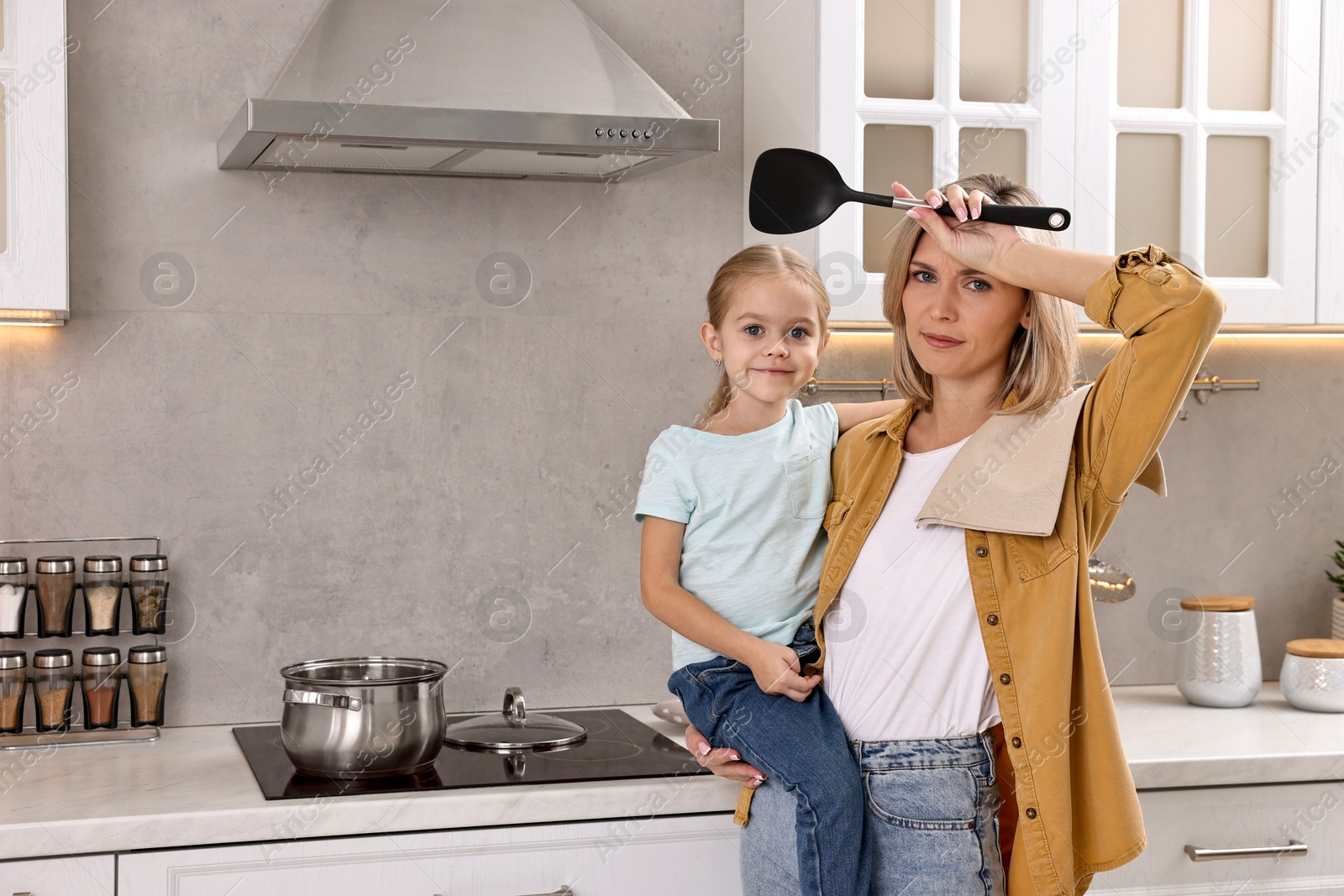 Photo of Tired housewife holding her daughter while cooking in kitchen