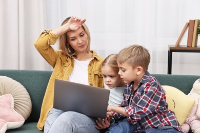 Photo of Tired housewife spending time with her kids and using laptop on couch at home