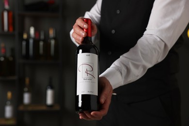 Photo of Professional sommelier holding bottle of red wine indoors, closeup