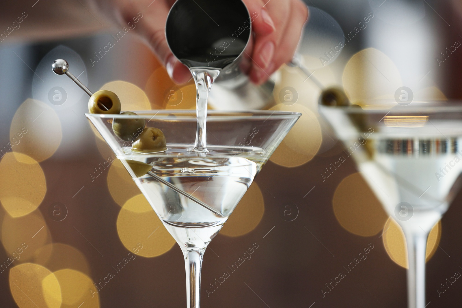 Photo of Woman making tasty martini cocktail with olives on blurred background, closeup. Bokeh effect