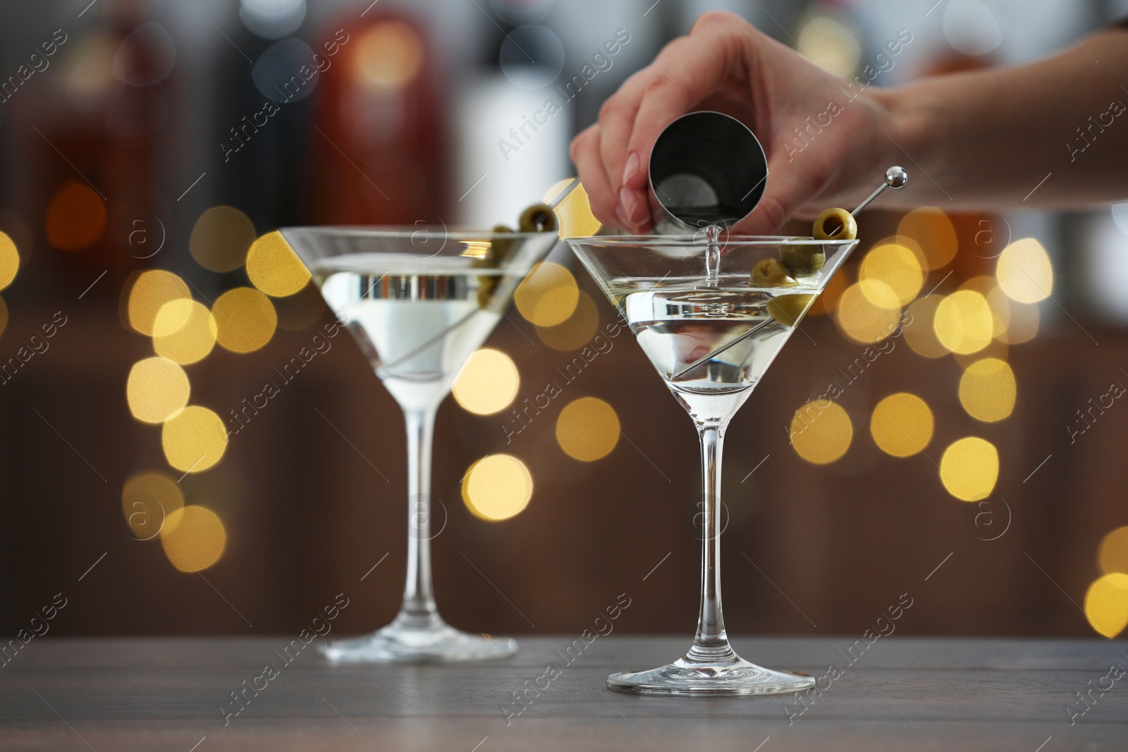 Photo of Woman making tasty martini cocktail with olives at wooden table on blurred background, closeup. Bokeh effect