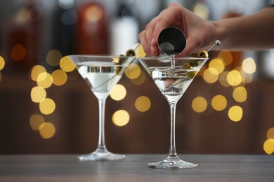 Photo of Woman making tasty martini cocktail with olives at wooden table on blurred background, closeup. Bokeh effect