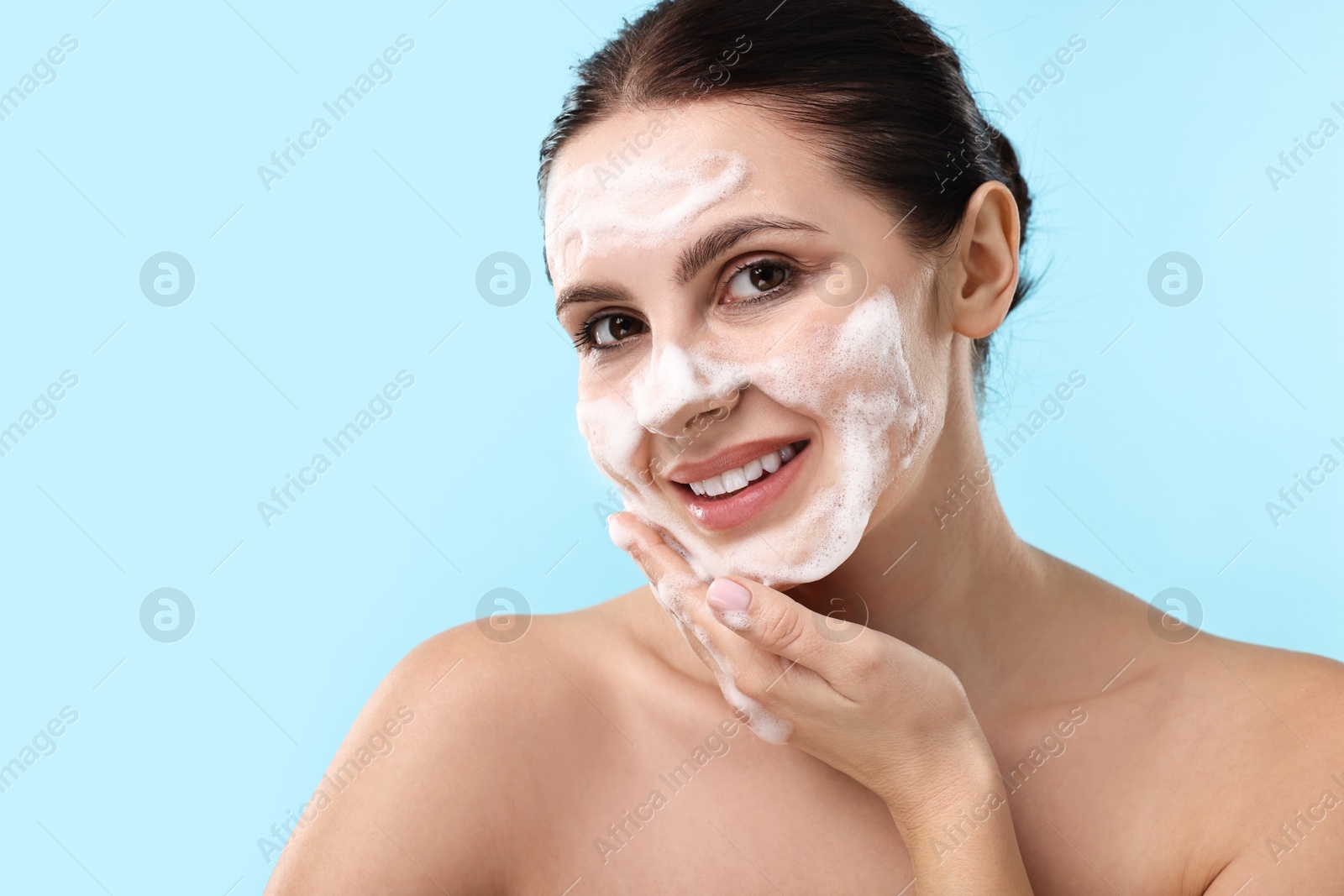 Photo of Smiling woman washing her face with cleansing foam on light blue background, space for text