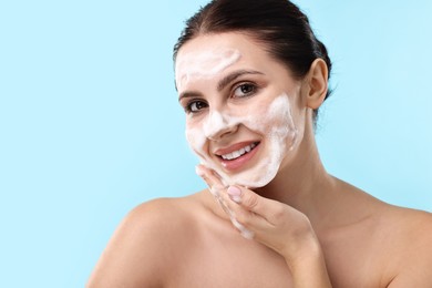 Smiling woman washing her face with cleansing foam on light blue background, space for text