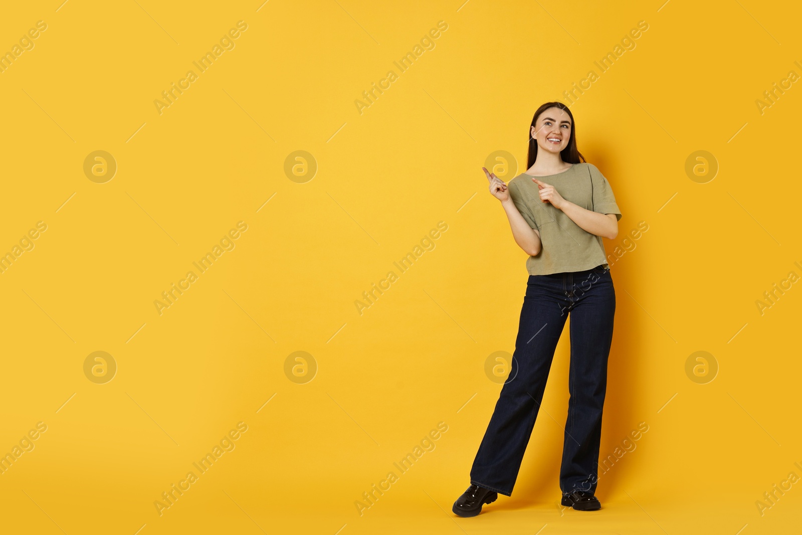 Photo of Smiling woman in stylish jeans pointing at something on orange background. Space for text