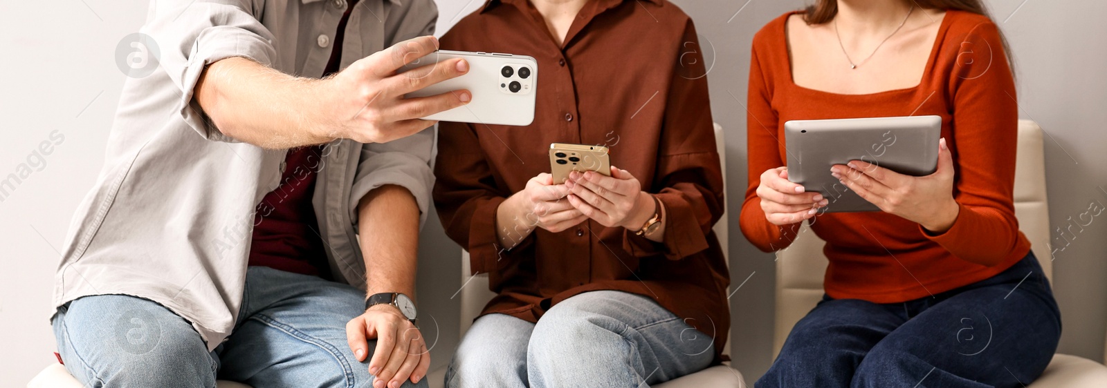 Photo of People using different gadgets on chairs near grey wall indoors, closeup. Modern technology