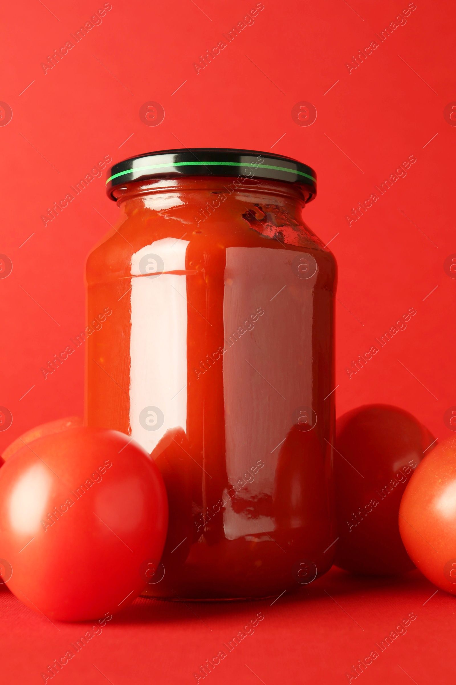 Photo of Ketchup in glass jar and fresh tomatoes on red background, closeup