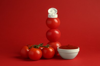 Photo of Stack of fresh tomatoes with plastic cap as bottle and ketchup in bowl on red background