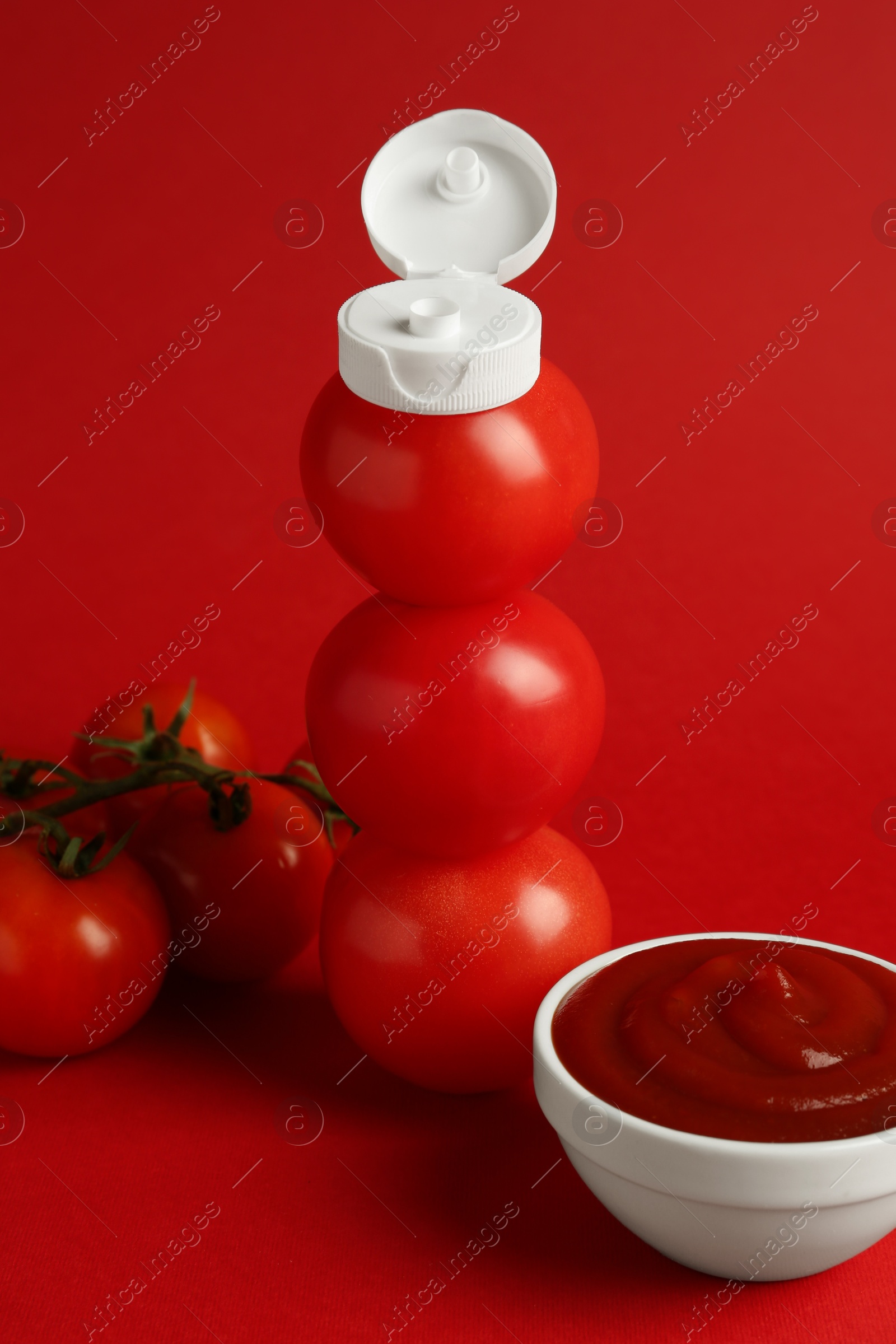 Photo of Stack of fresh tomatoes with plastic cap as bottle and ketchup in bowl on red background