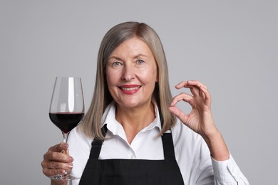 Senior woman with glass of wine showing okay gesture on grey background