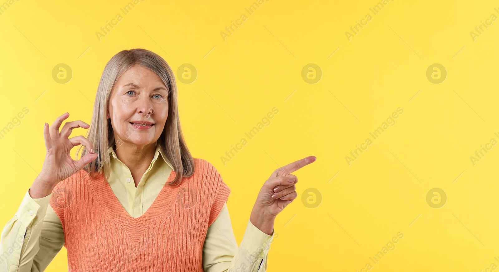 Photo of Senior woman showing okay gesture while pointing at something on yellow background. Space for text