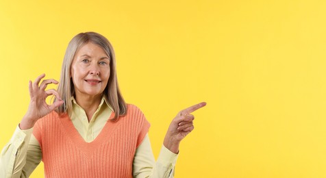 Photo of Senior woman showing okay gesture while pointing at something on yellow background. Space for text