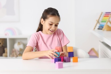 Photo of Happy girl building pyramid of colorful cubes at table indoors