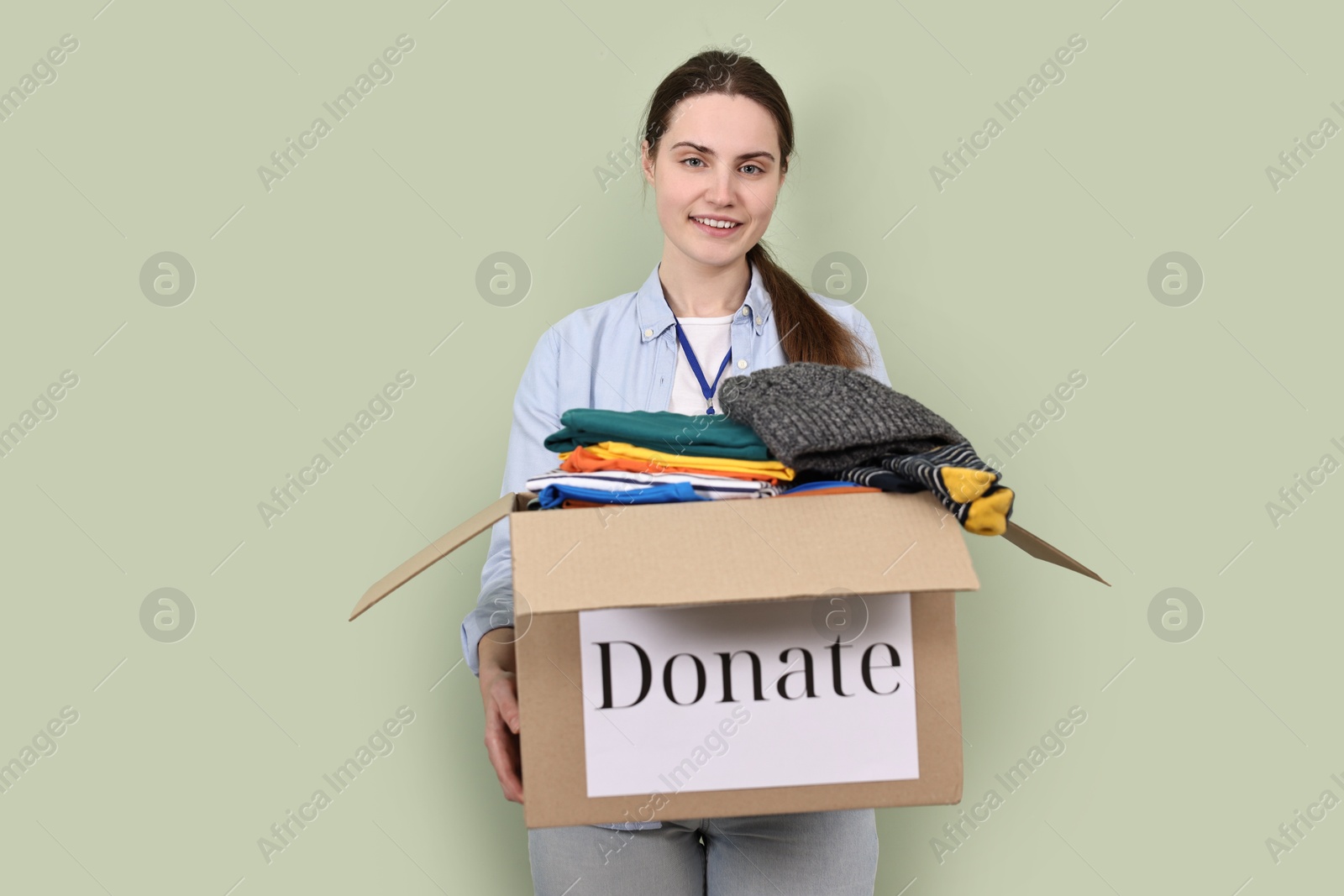 Photo of Woman holding donation box with clothes on pale olive background