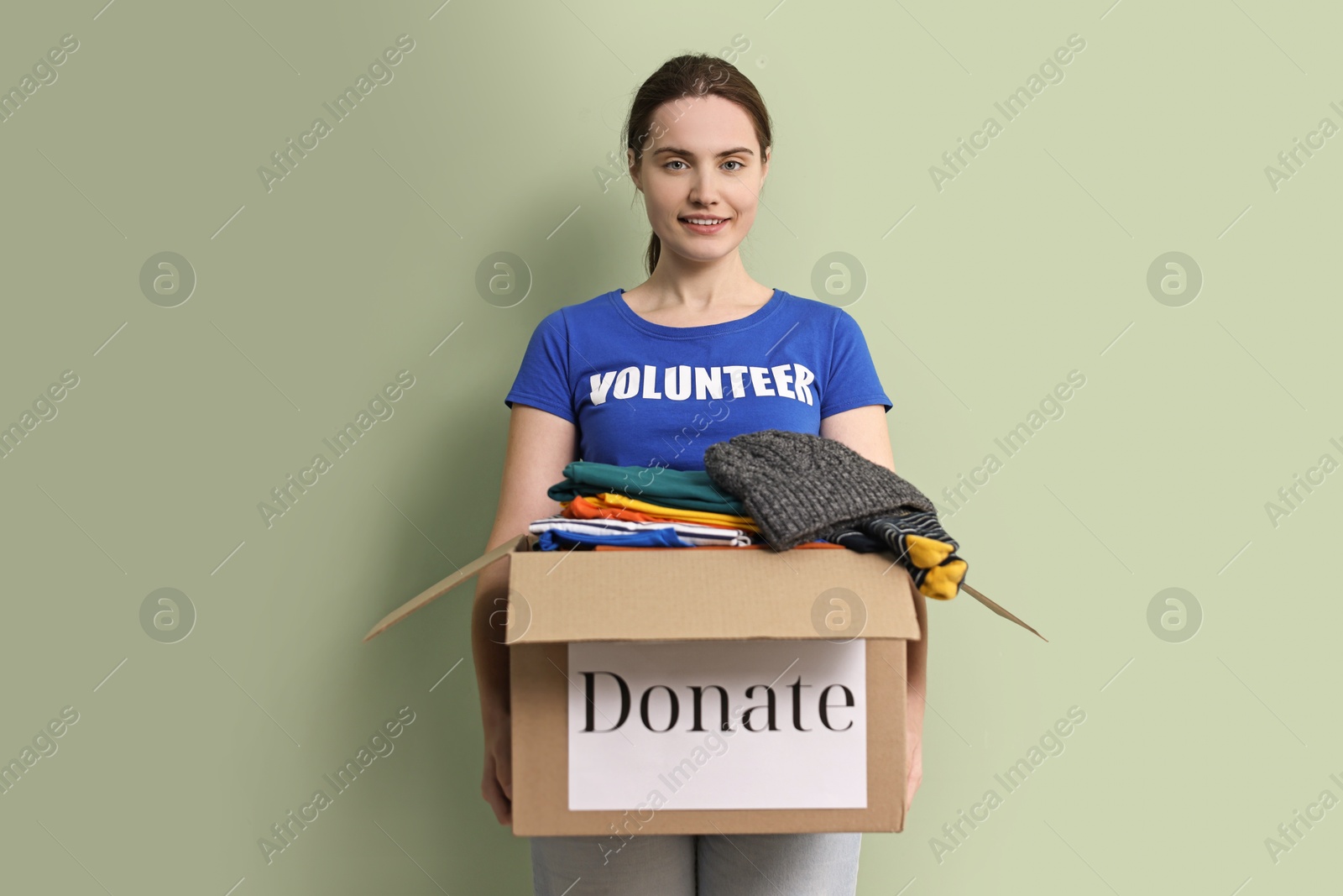 Photo of Woman holding donation box with clothes on pale olive background