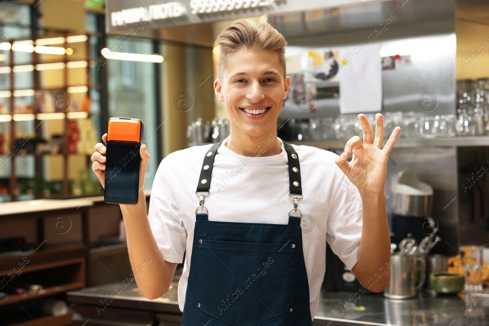 Photo of Smiling cafe worker with payment terminal showing OK gesture indoors