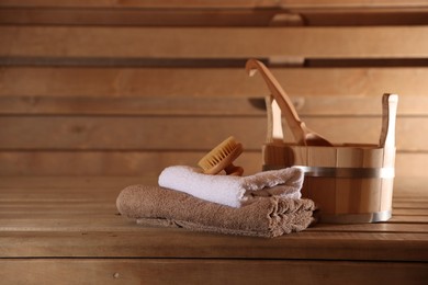 Photo of Bucket, ladle, massaging brush and stack of towels on wooden bench in sauna. Space for text