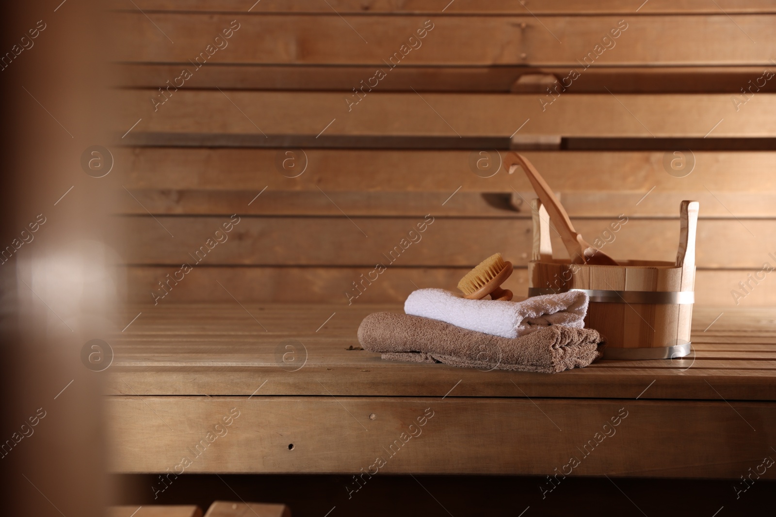 Photo of Bucket, ladle, massaging brush and stack of towels on wooden bench in sauna. Space for text