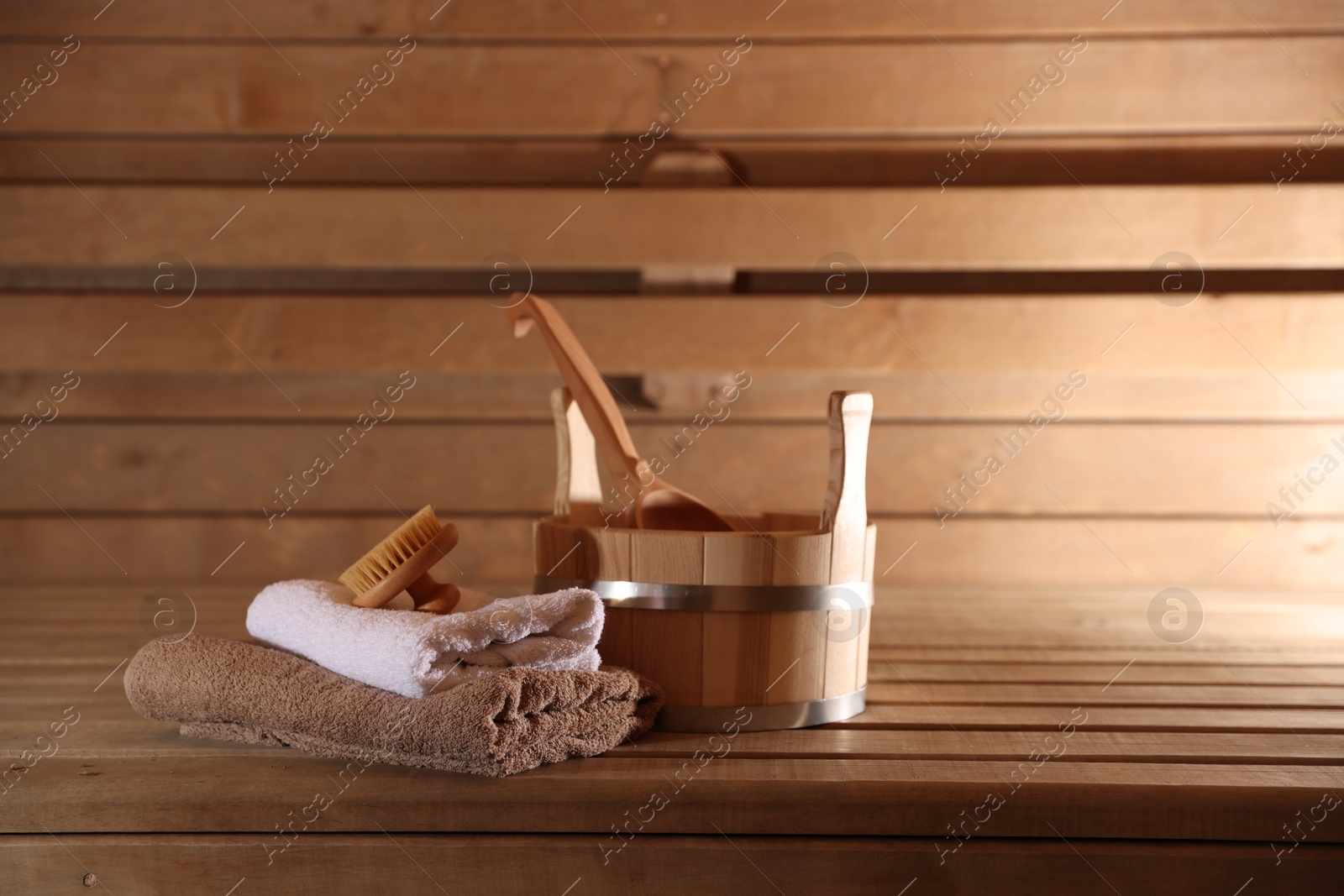 Photo of Bucket, ladle, massaging brush and stack of towels on wooden bench in sauna. Space for text