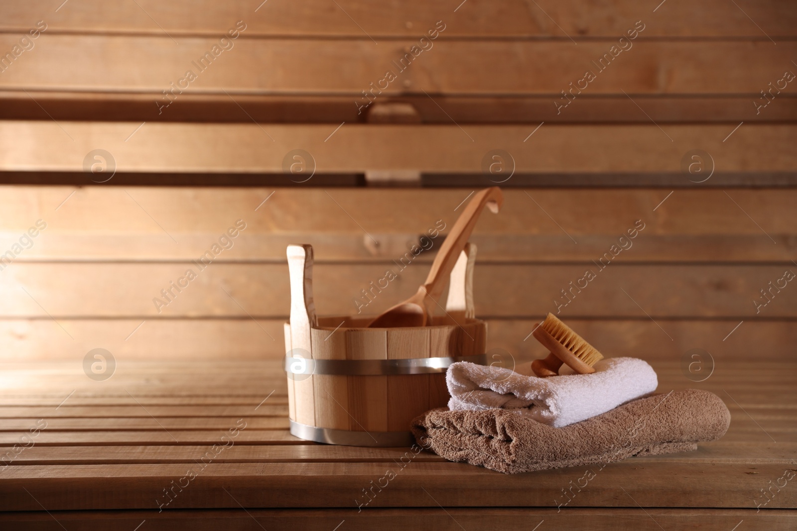 Photo of Bucket, ladle, massaging brush and stack of towels on wooden bench in sauna. Space for text