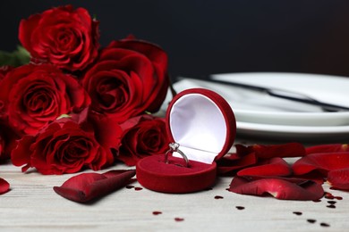 Photo of Engagement ring in box, roses, confetti and tableware on light wooden table, closeup. Romantic dinner