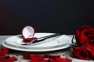 Photo of Beautiful setting for romantic dinner and engagement ring surrounded by roses and confetti on light table, closeup