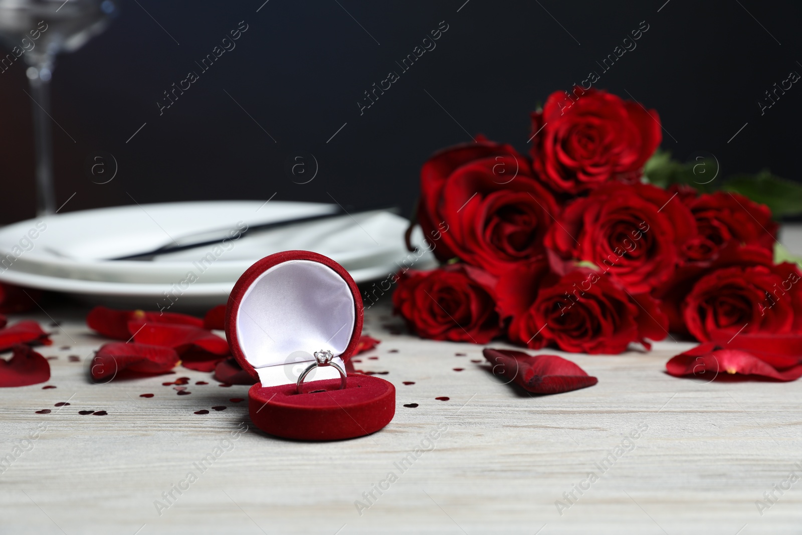 Photo of Engagement ring in box, roses, confetti and tableware on light wooden table, closeup. Romantic dinner